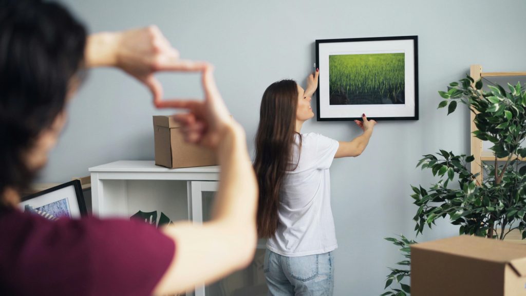 woman hanging a picture of nature on the wall while a man gestures with his fingers to frame the placement