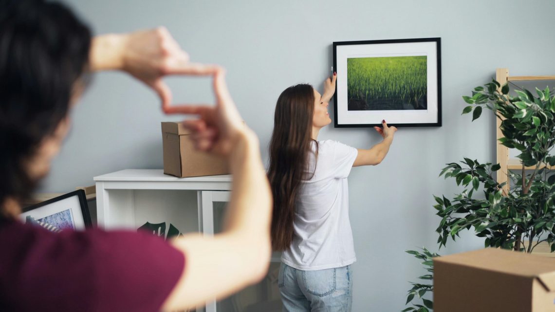 woman hanging a picture of nature on the wall while a man gestures with his fingers to frame the placement