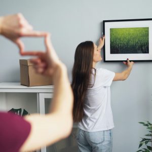 woman hanging a picture of nature on the wall while a man gestures with his fingers to frame the placement