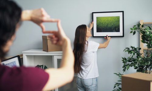 woman hanging a picture of nature on the wall while a man gestures with his fingers to frame the placement
