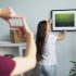 woman hanging a picture of nature on the wall while a man gestures with his fingers to frame the placement