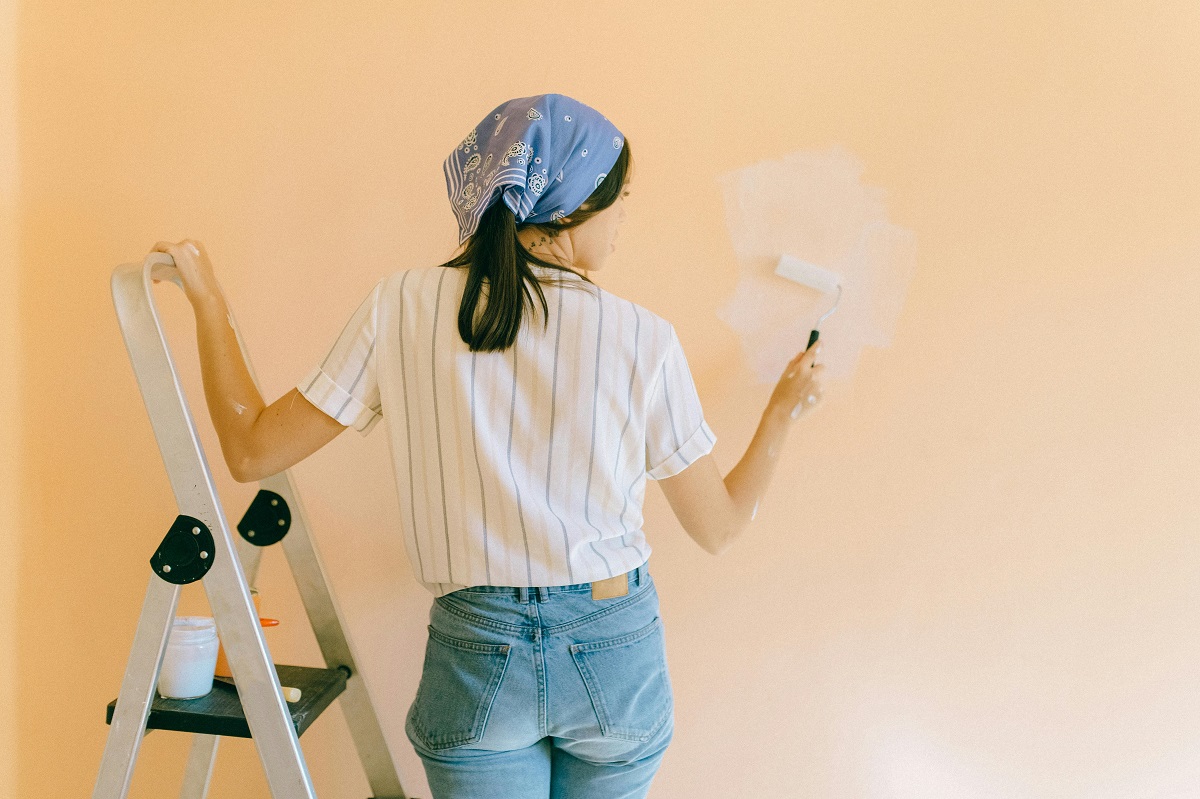 woman standing on a ladder, painting an exterior wall yellow with a paint roller